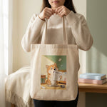 Homework Eaten Pet Tote Bag. A person holding the natural canvas tote bag in front of them with both hands, cropped from the chin down, cream wall behind.