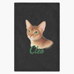 Colored pencil portrait of a ginger Abyssinian cat printed on a charcoal gray fleece blanket, with the name "Cleo" drawn below.