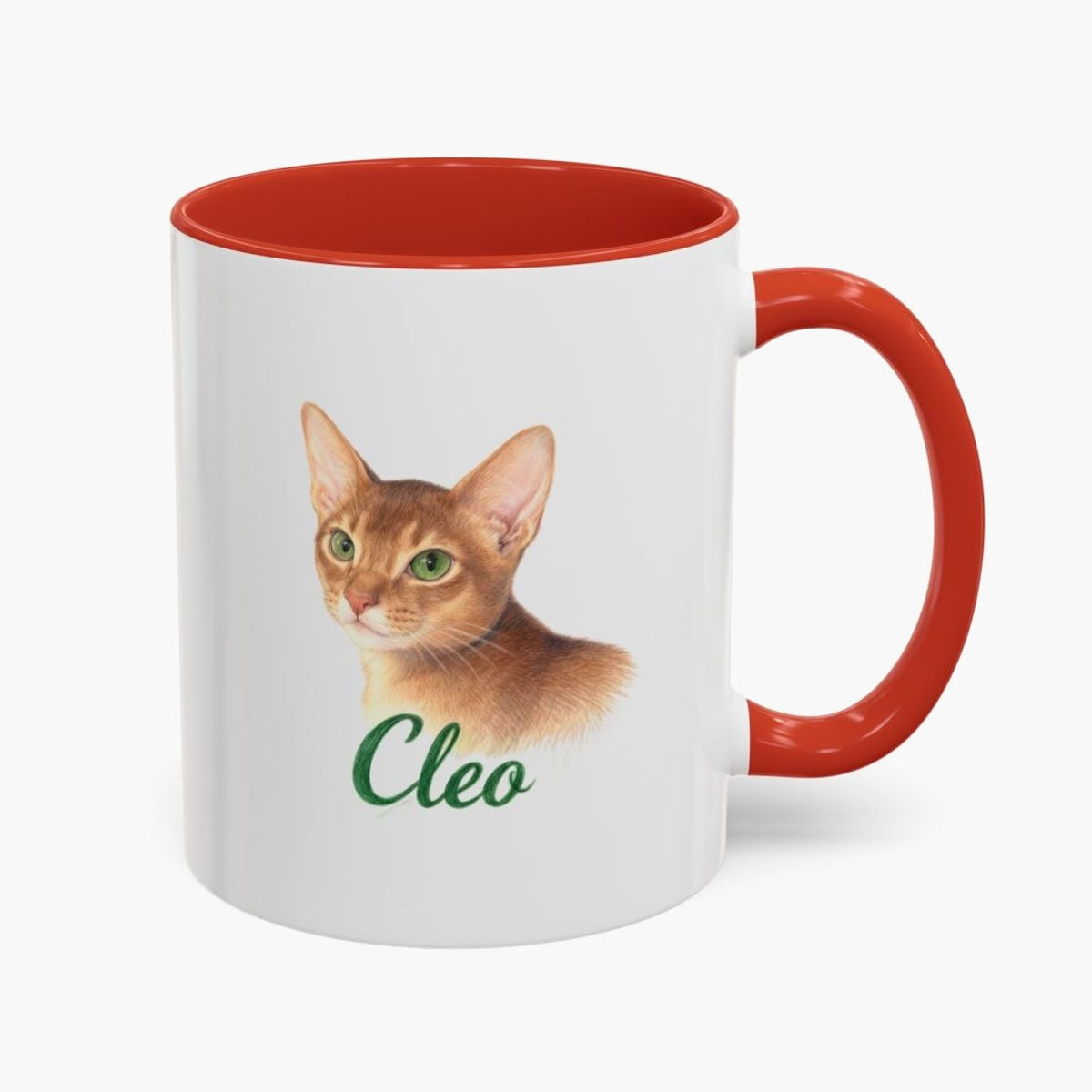 Colored pencil portrait of a ginger Abyssinian cat on a 11 oz mug with a red handle, with the name "Cleo" drawn below.