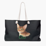 Colored pencil portrait of a ginger Abyssinian cat on a charcoal gray weekender bag with white rope handles, with the name "Cleo" drawn below.