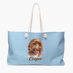 Colored pencil portrait of an apricot labradoodle on a french blue weekender bag with white rope handles, with the name "Cooper" drawn below.