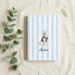 Student Watercolor Pet Notebook. Overhead flat lay of the hardcover notebook on cream linen with a sprig of eucalyptus and a single brass paperclip.