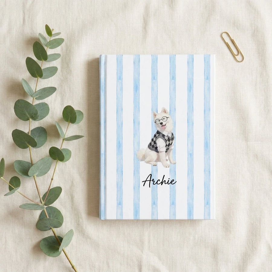 Student Watercolor Pet Notebook. Overhead flat lay of the hardcover notebook on cream linen with a sprig of eucalyptus and a single brass paperclip.