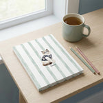 Student Watercolor Pet Notebook. The hardcover notebook lying on a pale oak desk beside a window, a sage ceramic mug and two sharpened pencils nearby.