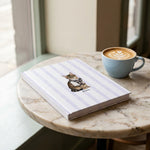 Student Watercolor Pet Notebook. The hardcover notebook on a small marble cafe table next to a latte in a pale blue cup.