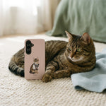 Student Watercolor Pet Phone Case. A real brown tabby cat with green eyes lying on a cream rug in soft daylight with the phone case resting on the floor near its paws.