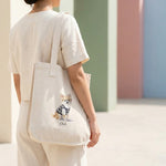 Student Watercolor Pet Tote Bag. A person walking away from the camera with the natural canvas tote bag over one shoulder, cream outfit, soft daylight, no face visible.