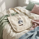Student Watercolor Pet Tote Bag. The natural canvas tote bag resting on rumpled cream bedding in soft morning light.